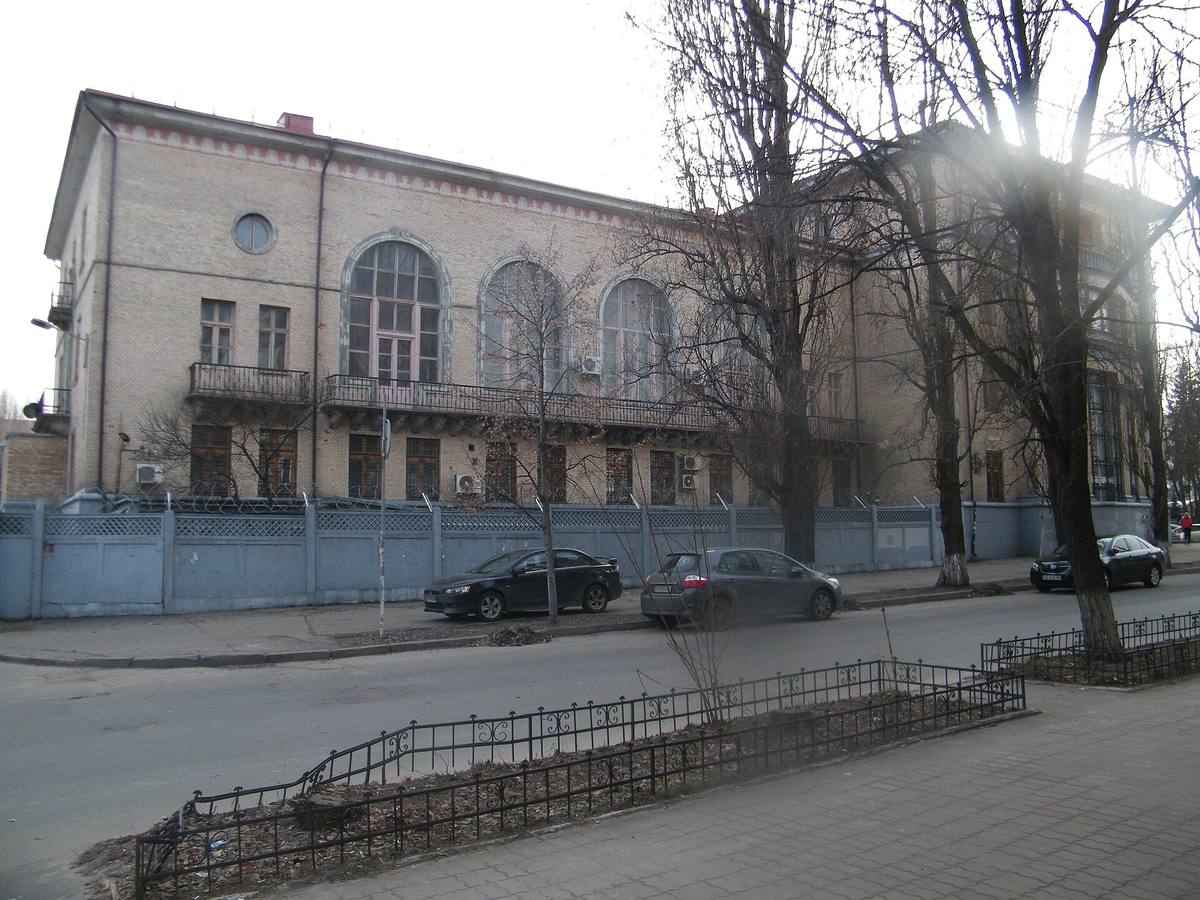 Dvoupatrová cihlová budova s obloukovými okny za modrým plotem, před ní ulice se zaparkovanými auty