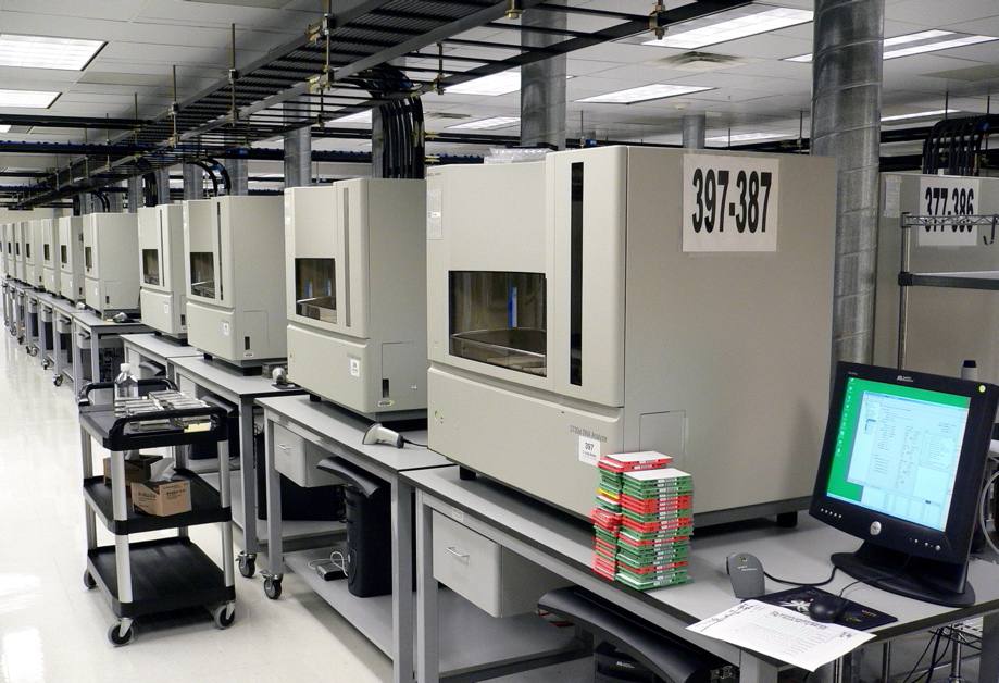 Dlouhá řada béžových sekvenátorů DNA na stolech v laboratoři, vpředu monitor a krabičky s destičkami