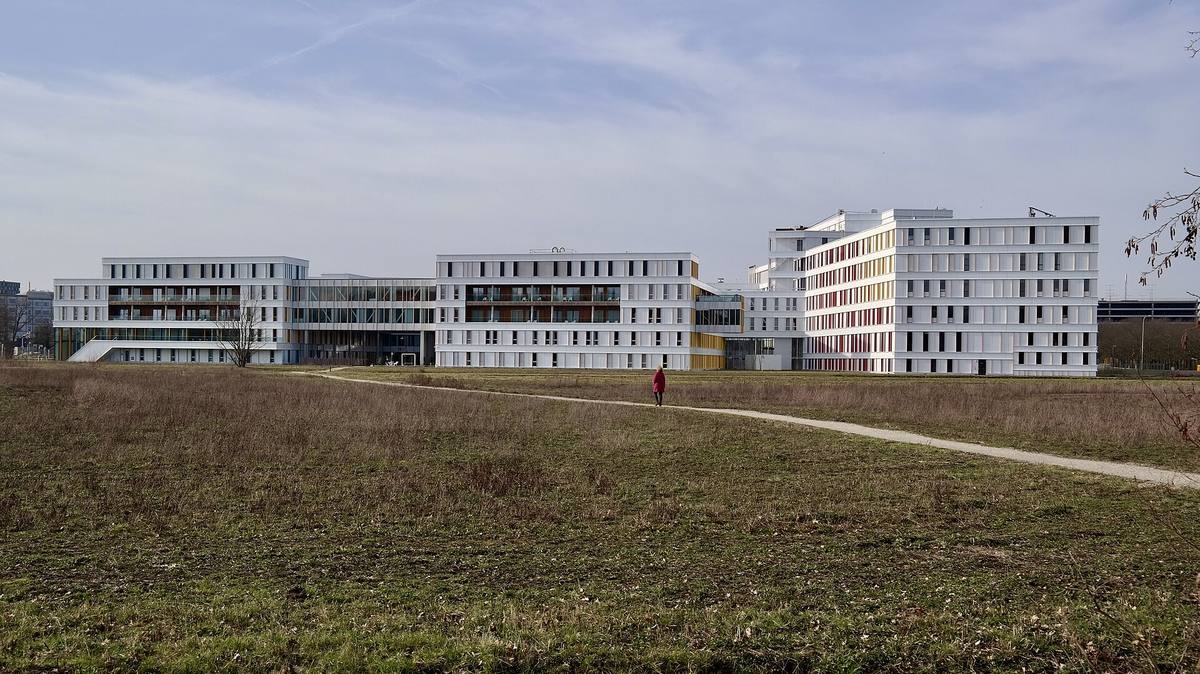 Nízké bílé budovy dětského onkologického centra na okraji města, v popředí travnatá pláň s pěšinou