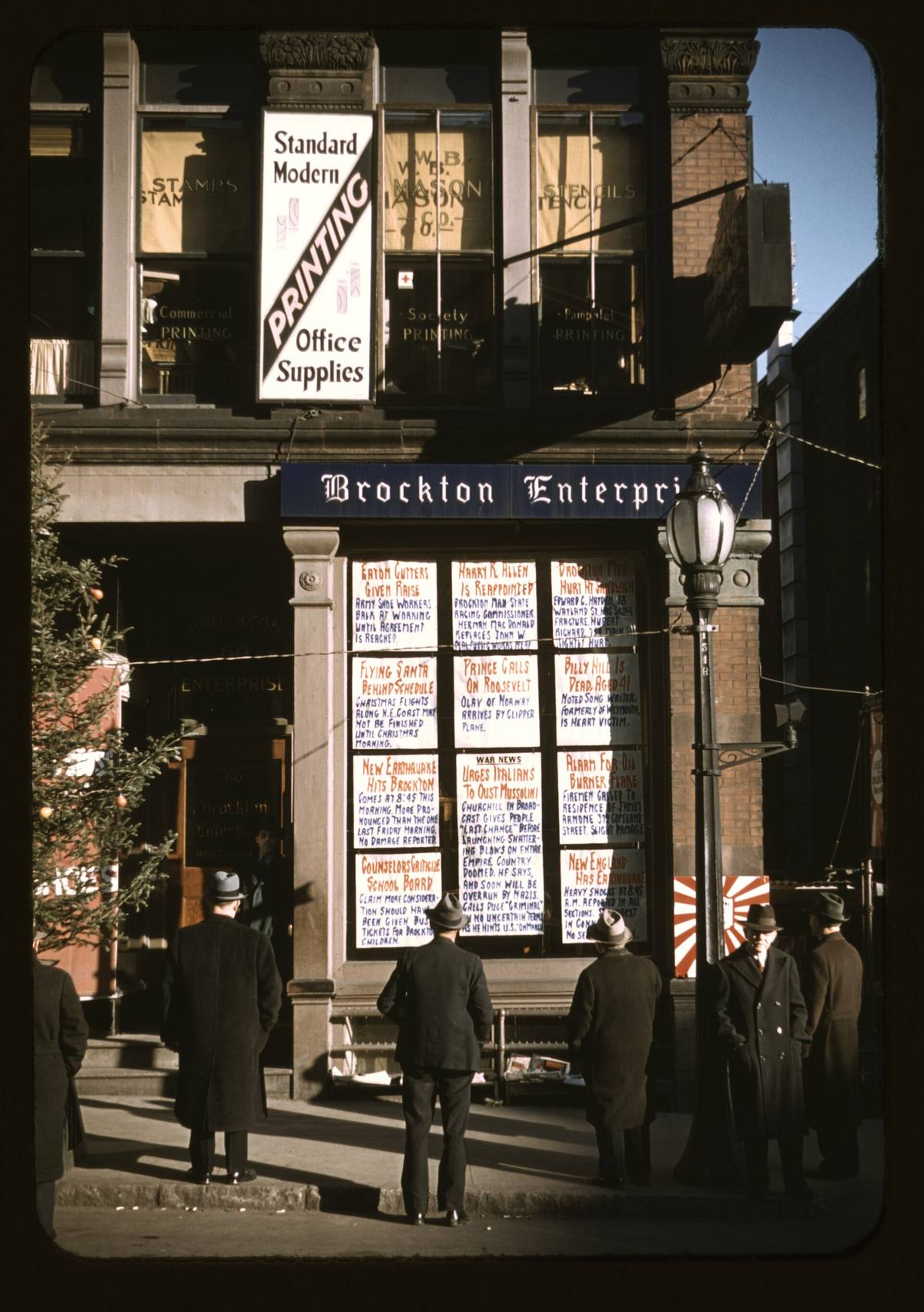 Skupina mužů čte novinové titulky vyvěšené ve výloze redakce, barevná fotografie z roku 1940