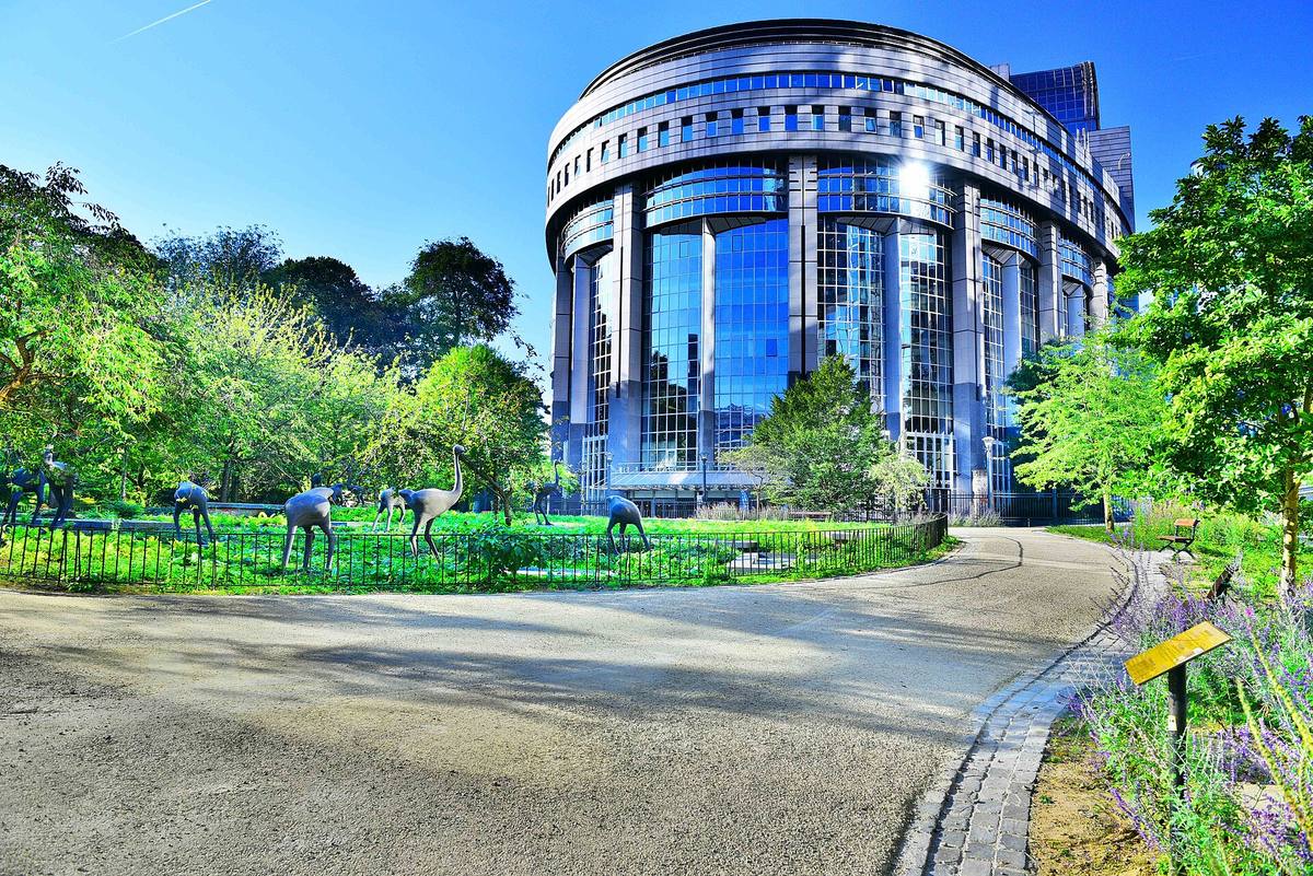 Zaoblená prosklená budova Evropského parlamentu v Bruselu, v popředí park se sochami
