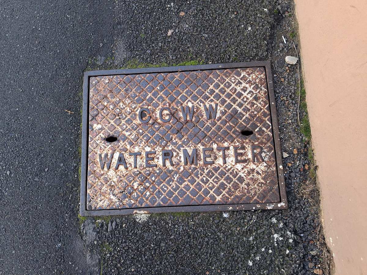 Litinový kryt šachty s vodoměrem v chodníku, na krytu nápis CCWW Water Meter